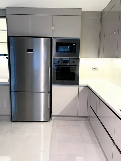 A clean and contemporary L-shaped kitchen in Gurgaon, showcasing seamless integration of a refrigerator and built-in oven tower. The high-gloss grey cabinetry and handle-less design create a sleek, minimalist aesthetic.
