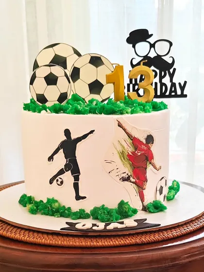 An "Official Teenager" football cake for a 13th birthday. The design features player silhouettes and soccer ball toppers on a bed of green buttercream "grass."