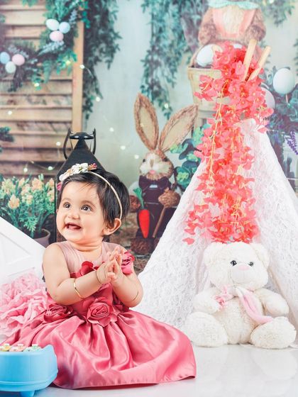 A beautiful baby girl in a pink dress, enjoying a whimsical garden party setup with a teddy bear and a teepee.