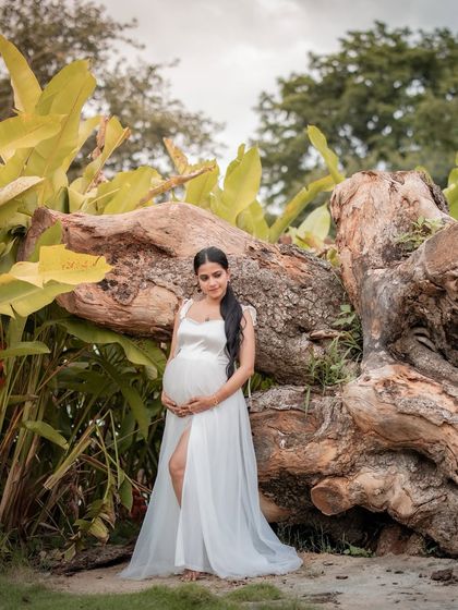 A serene moment in nature, with the mother-to-be cradling her bump in a simple white gown, standing by the roots of a large tree.