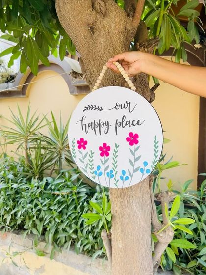 Another look at our 'Our Happy Place' wooden sign, hanging on a tree. The hand-doodled flowers and calligraphy make this a charming and welcoming piece for any home.
