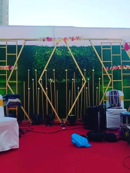 A geometric stage backdrop for a modern wedding reception. This outdoor setup uses a green hedge wall with intersecting gold diamond frames, accented with small floral arrangements and candle-like lights.