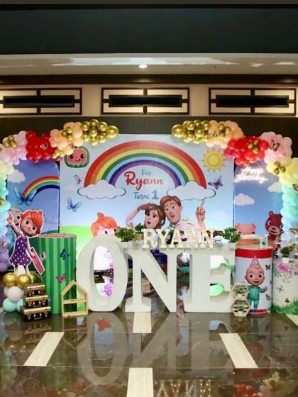 A grand Cocomelon setup with a rainbow backdrop. The stage features large 'ONE' letters, character cutouts, and themed plinths, creating a vibrant and exciting party scene.