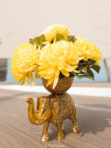 A charming decor detail for a Haldi ceremony. A small, ornate golden elephant statue holds a bouquet of bright yellow chrysanthemums.