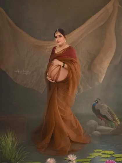 This standing portrait showcases a modern interpretation of a traditional saree drape, using sheer brown fabric to highlight the baby bump. The peacock and lotus pond in the background add to the painterly, culturally rich feel of this artistic maternity photo.