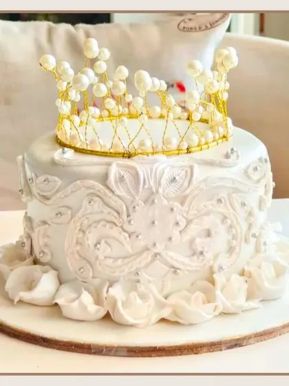 A regal white cake with intricate lace detailing, topped with a delicate, handcrafted pearl and gold crown.