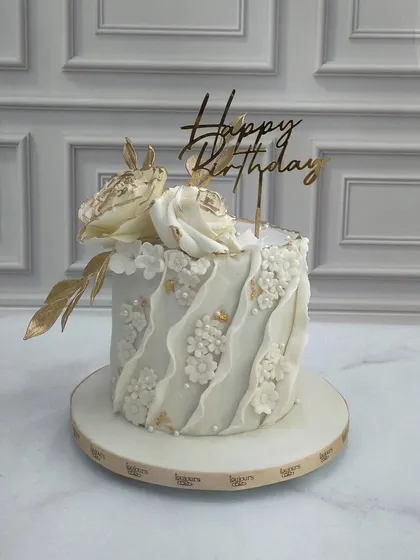 An elegant and textured birthday cake in white and gold. This design features a delicate fabric-like drape effect, tiny floral details, and is topped with two beautiful white sugar roses brushed with edible gold.