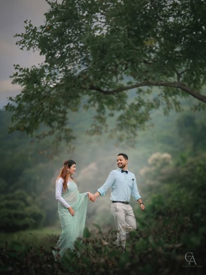A couple walks hand in hand under the branch of a large tree in a foggy, green landscape. This image captures a simple, happy moment that feels timeless and natural.