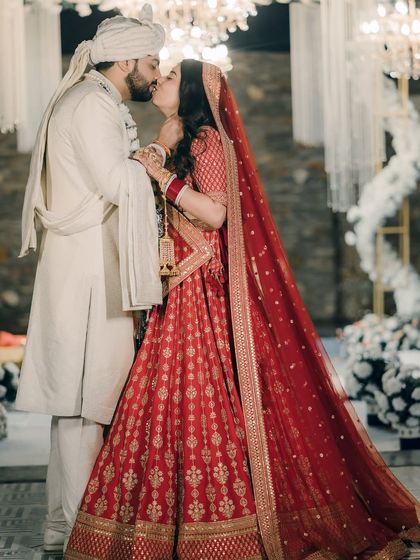 A timeless wedding day kiss, with the bride in her vibrant red lehenga and the groom in an elegant white sherwani. This classic shot captures the pinnacle of their celebration against a beautifully decorated backdrop.