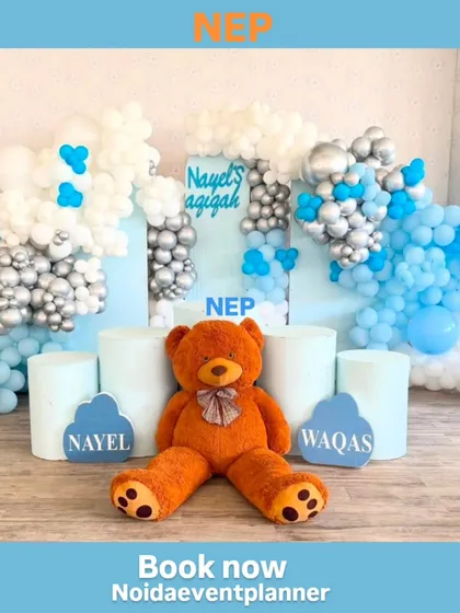 The main photo booth for an Aqiqah ceremony, featuring a large teddy bear, personalized name cutouts, and a backdrop with silver, white, and blue balloons.