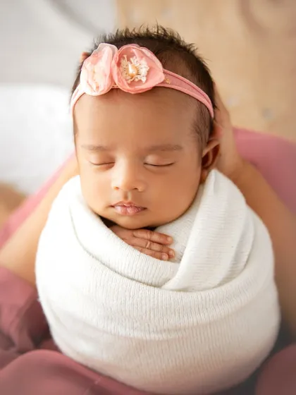 A close-up portrait of a sleeping newborn, tightly swaddled to keep her calm and content. The floral headband adds a soft, delicate touch.
