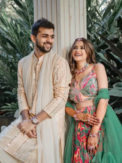 A beautiful, candid moment from a Haldi ceremony. The couple shares a genuine laugh, their colorful outfits vibrant against the green background, showcasing the joy of wedding festivities.