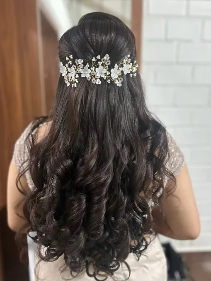 A perfect hairstyle for a reception or engagement. This is a classic half-up style with voluminous, bouncy curls and a delicate floral hair accessory that adds a touch of modern elegance.