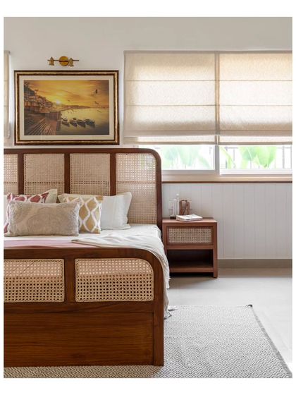 A close-up of the teakwood and cane bed, set against pristine white paneled walls. A gold picture light illuminates the Varanasi painting, enhancing the room's nostalgic and serene feel.
