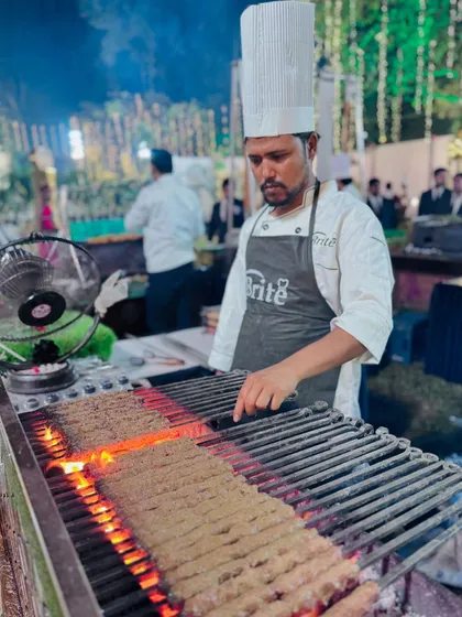 A chef at the live grill station, expertly turning Seekh Kebabs. The fan in the background helps maintain the perfect coal temperature for grilling.