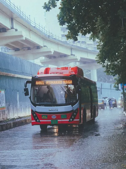 A red electric bus navigates the wet streets of Powai. The rain adds a dramatic flair to everyday city life, especially with the flyover architecture in the background.
