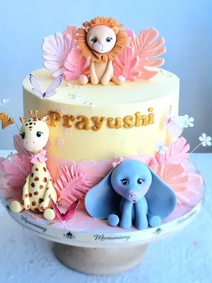 A two-tier safari cake with a soft, creamy yellow finish and pink accents. This design features a lion, giraffe, and elephant, all handmade from fondant, creating a sweet and gentle theme for a young child's birthday.