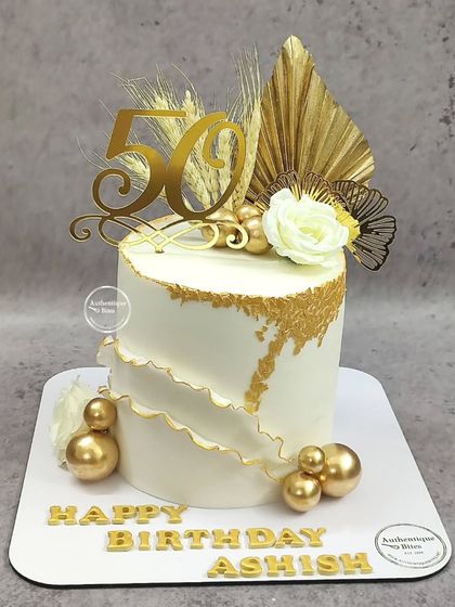 A chic white and gold 50th birthday cake. This design features a textured wave effect, gold spheres, and dried botanicals for a modern, celebratory look.
