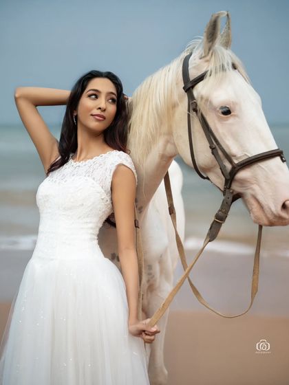 A portrait of the model looking back while standing with the horse. The soft evening light and the serene ocean background create a peaceful and elegant fashion photograph.