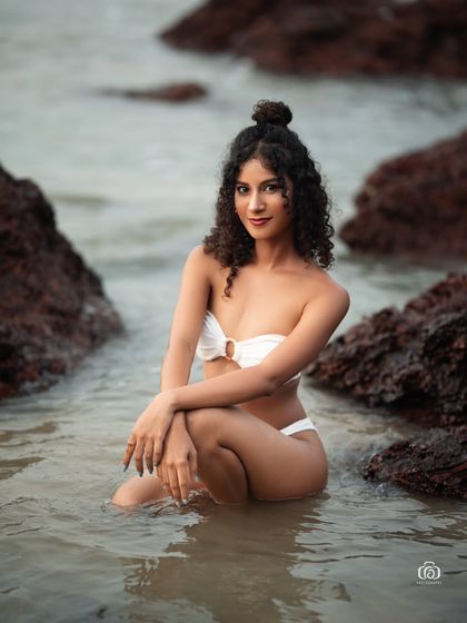A serene portrait of a model sitting in the shallow water between the rocks. The calm water and soft light create a peaceful and beautiful beach scene.