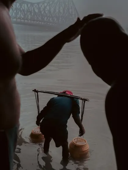 A powerful silhouette of men at the Hooghly river, with the Howrah Bridge in the distance. The composition focuses on the human form and their connection to the water.