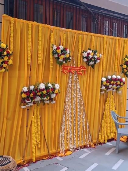 A simple yet beautiful Haldi backdrop for a home event. The bright yellow fabric is decorated with marigold garlands and small bouquets of red, white, and yellow flowers.