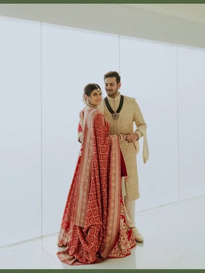 A full-length portrait against a clean, minimalist background. The bride's stunning red lehenga flows beautifully as she stands with her groom, creating a modern and elegant wedding photo.