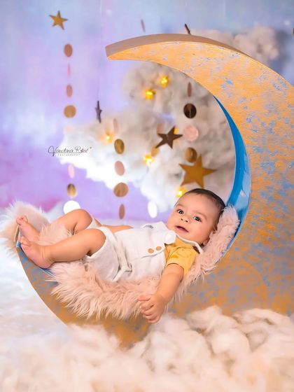 A dreamy 'over the moon' theme for a baby photoshoot. The baby relaxes on a crescent moon prop surrounded by fluffy clouds and hanging stars, creating a magical celestial scene.