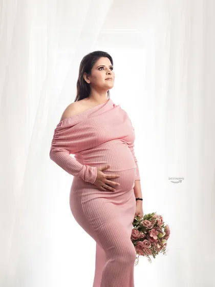 A classic three-quarter maternity portrait. The client looks radiant in a pink off-shoulder gown, holding a matching bouquet against a bright and airy studio background.