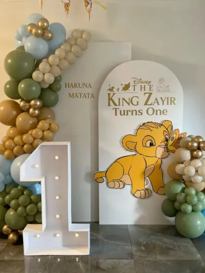 Hakuna Matata! A sweet Lion King theme for a first birthday celebration. This setup includes a baby Simba cutout, a marquee number "1", and a balloon garland in gentle greens, blues, and browns.