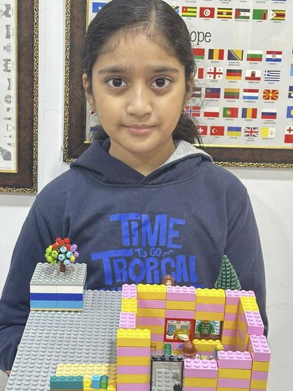 A girl displays the multi-level LEGO house she built, featuring pink and yellow walls and interior details.