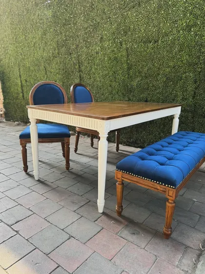 A classic dining set with a twist. It features a teak table with a white-painted base, two royal blue upholstered chairs, and a matching tufted bench for flexible seating.