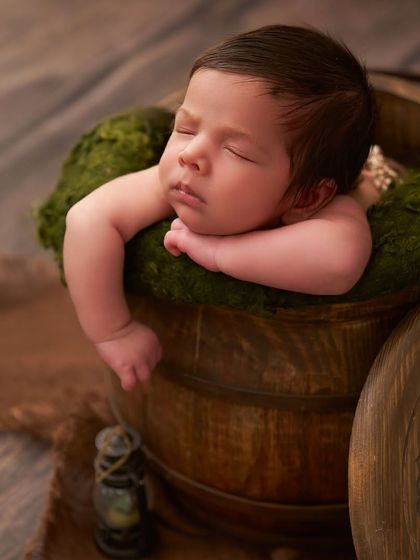 A close-up of a newborn's peaceful face as they rest their head on a mossy bed inside a rustic wooden bucket.