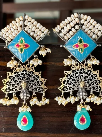 Traditional design meets fusion style in these stunning earrings. They feature a pearl-studded top with a light blue enamel base, leading to a beautifully detailed silver and gold-plated peacock with dangling pearl and gemstone drops.