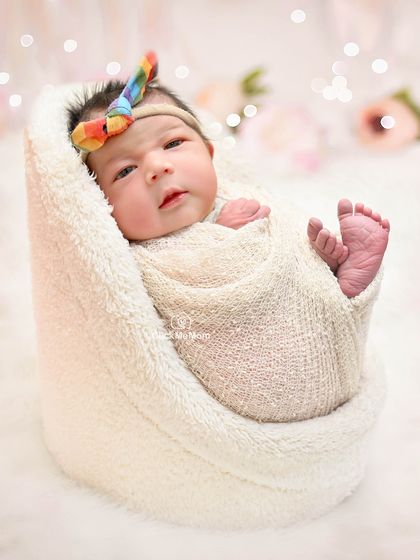 This baby girl is awake and looking at the camera, wrapped in a cozy white blanket with a rainbow bow adding a pop of color.