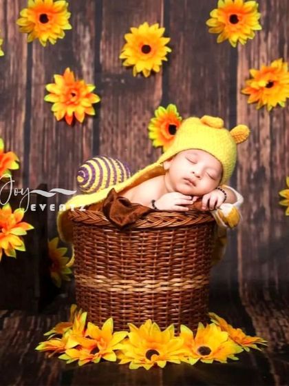 A sweet and cheerful honey bee theme for a newborn photoshoot. The baby wears a yellow knit hat and is nestled in a wicker basket against a backdrop of sunflowers and wood panels.