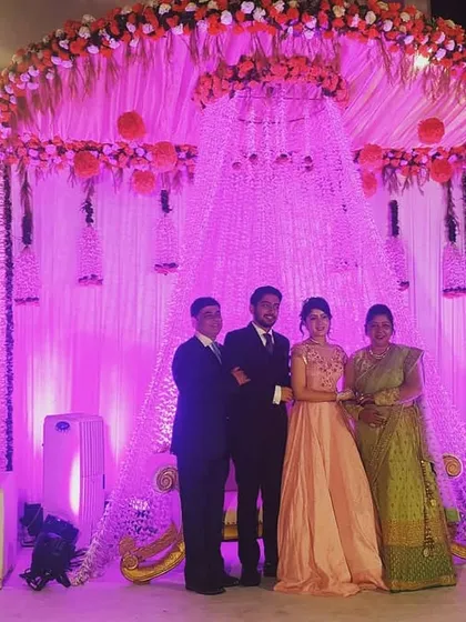 A family poses on the reception stage we designed. The backdrop features a cascade of floral strings and hanging lanterns under a canopy of pink flowers, all illuminated by soft purple lighting.
