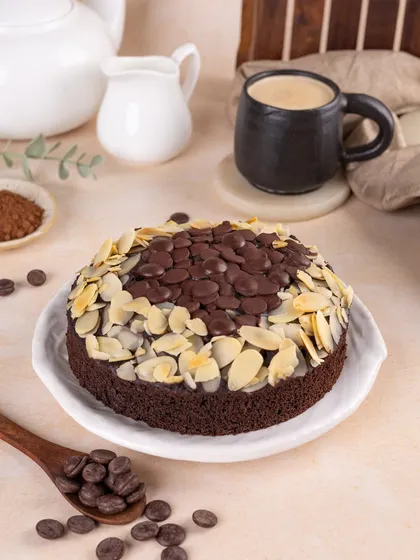 A full view of the beautiful chocolate and almond cake. The styling with a coffee pot and scattered chocolate chips tells a story of a relaxing coffee break.