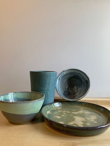 A collection of my handmade tableware, including a tall tumbler, bowls, and a speckled plate. The earthy green and blue glazes create a cohesive, calming set.
