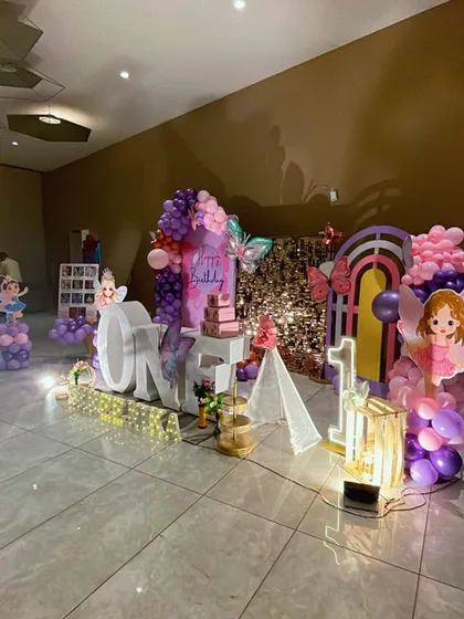 A whimsical butterfly and fairy themed first birthday party. This setup features large 'ONE' marquee letters, a sparkling backdrop, and beautiful fairy cutouts, all surrounded by purple and pink balloon decorations.