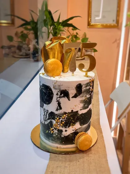 A modern and artistic cake for a 75th birthday. This tall cake features a black and white abstract painted design with gold leaf and pearl sprinkles, topped with a gold sail and macarons.