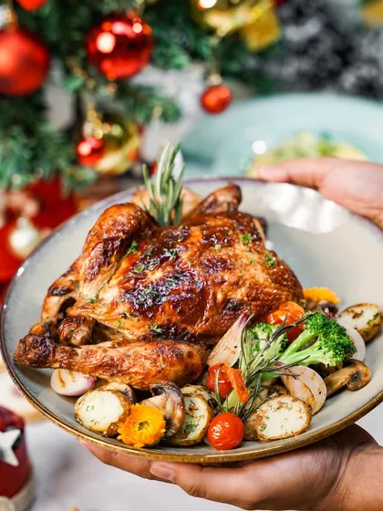 Hands presenting a beautifully roasted whole chicken for a Christmas meal. The warm, festive background and focus on the main course make this an inviting and celebratory shot.
