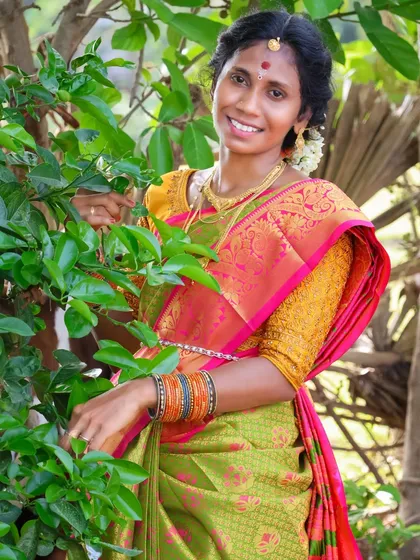 A serene portrait taken in a natural setting. The woman is framed by green leaves, which complements her saree and creates a fresh, organic feel for this traditional photoshoot.