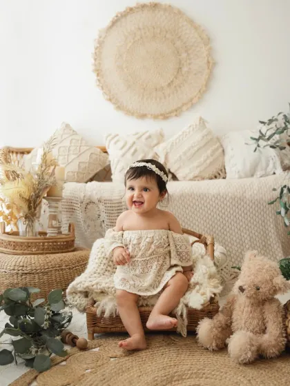 A happy baby girl sits in a wicker basket, surrounded by bohemian decor. This is a lovely setup for a sitter session or milestone photoshoot.