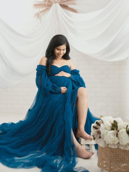 A serene and gentle portrait. The mom-to-be is seated, cradling her bump in a beautiful blue gown, with soft white drapes and flowers creating a peaceful atmosphere.