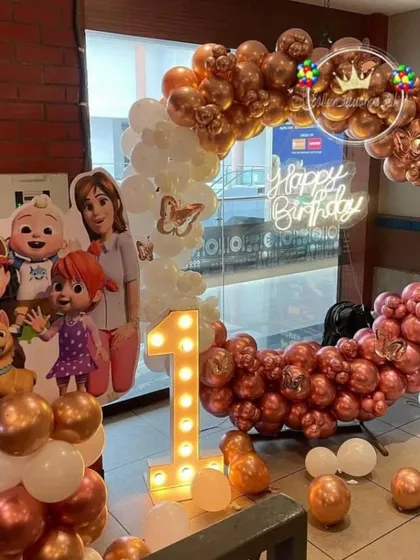 A Cocomelon first birthday theme in rose gold and white. This setup includes a balloon ring, a large character family cutout, and a light-up number '1'.