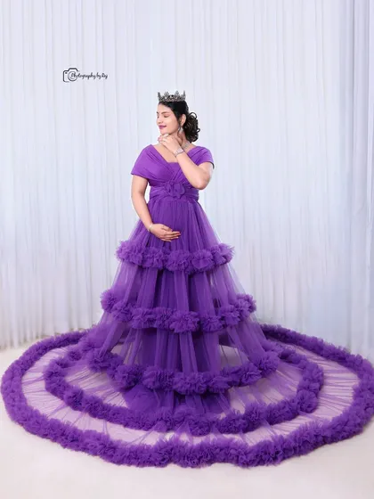A regal studio portrait of a mother-to-be in a purple ruffled gown and crown, with the dress spread out beautifully.