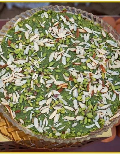 A traditional Indian green sweet, possibly a pistachio barfi, generously topped with sliced almonds and pistachios. A classic dessert that's rich in flavor.