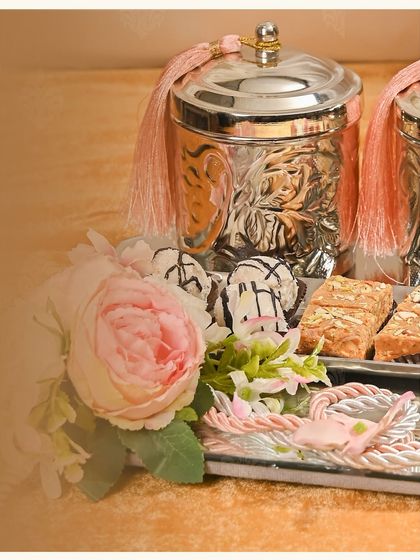 From my Miraaya Wedding Edit, this hamper is an ode to timeless grace. It features silver-plated jars with pink tassels and a selection of gourmet sweets, all arranged with beautiful flowers.