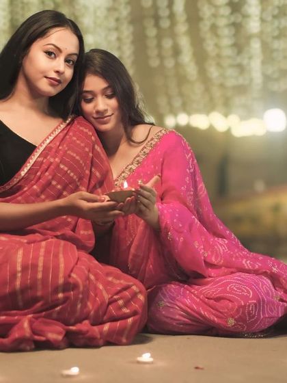 A tender moment of friendship during Dev Deepawali, as two women in sarees share the light of a diya against a backdrop of festive lights.